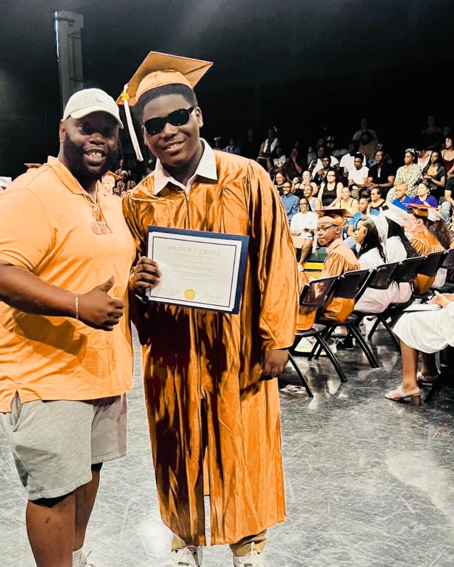 A mentor gives a thumbs-up beside a smiling graduate in cap and gown holding his diploma